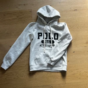 Grå hoodie från Polo Ralph Lauren - Snygg ljusgrå hoodie från Polo Ralph Lauren med svart tryck framtill där det står 'POLO 1967 ATHL. DEPT.'. Tröjan har huva med snörning och en stor magficka. Perfekt för en chill och sportig stil.
