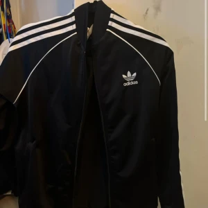 Svart Adidas track kofta med vita ränder. - Klassisk svart Adidas track kofta med vita ränder längs axlarna och ärmarna. Passar storlek 170 Cm nästan ny skick.
