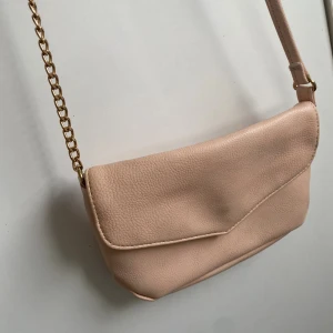Ljusrosa  axelväska med guldkedja - Snygg ljusrosa axelväska i syntetmaterial med guldfärgad kedjedetalj på axelremmen. Väskan har ett stilrent kuvertformat lock och flera praktiska fack inuti. Perfekt storlek för det viktigaste och passar till många olika outfits.