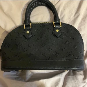 Svart väska  - Snygg svart handväska med klassiskt monogrammönster. Väskan har guldfärgade metalldetaljer och dragkedja upptill. Perfekt storlek för det viktigaste och en ikonisk accessoar till din outfit.