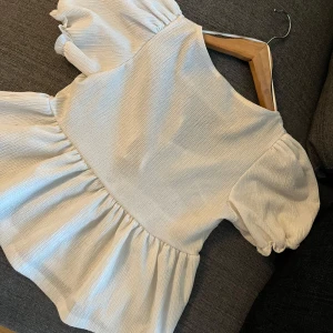 Vit blus med puffärm från Gina Tricot - Supersöt vit blus från Gina Tricot i storlek XS. Modellen har puffiga korta ärmar, volang nedtill och två knytband framtill. Tyget är lätt strukturerat och ger en trendig look. Perfekt för dig som gillar romantiska och feminina detaljer.