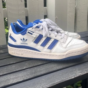 Adidas sneakers vit/blå retrostil - Säljer ett par klassiska Adidas sneakers i vitt och blått med de ikoniska tre ränderna på sidan. Skorna har snörning, rund tå och är tillverkade i skinn med blå textildetaljer. Perfekt för dig som gillar retrovibbar och streetstyle.