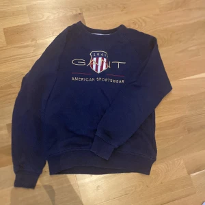 Marinblå sweatshirt från GANT - Säljer en marinblå sweatshirt från GANT med broderad logga och sköld på bröstet. Tröjan har rund halsringning, långa ärmar och ribbade muddar. Perfekt för dig som gillar klassisk och sportig stil.