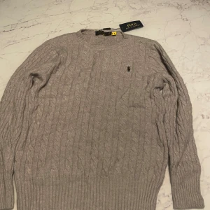 Ljusgrå kabelstickad tröja Polo Ralph Lauren - Kabelstickad ljusgrå tröja från Polo Ralph Lauren med klassisk rund halsringning och broderad logga på bröstet. Tröjan är i mjuk ull och har långa ärmar samt ribbade muddar för en snygg passform.