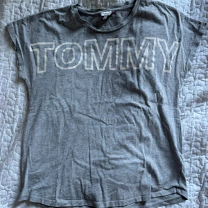 Grå t-shirt från Tommy Hilfiger - Snygg grå t-shirt från Tommy Hilfiger med stort tryck 'TOMMY' på framsidan och 'HILFIGER' på ryggen. Klassisk rund halsringning och korta ärmar. Tillverkad i mjuk bomull för en skön känsla hela dagen.
