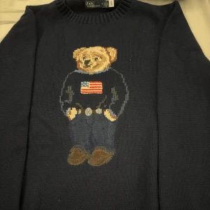 Mörkblå stickad Polo Bear-tröja - Säljer en ikonisk mörkblå stickad tröja från Polo Ralph Lauren med den klassiska Polo Bear broderad framtill. Tröjan har rund halsringning och långa ärmar. Perfekt statement-piece för dig som gillar preppy stil och vill sticka ut.