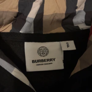 Svart Burberry hoodiejacka med dragkedja - Svart hoodiejacka från Burberry med klassisk rutig huva i beige, rött och svart. Jackan har dragkedja i guld, snörning i huvan och känguruficka framtill. Perfekt för dig som vill ha en stilren och lyxig streetwear-look.