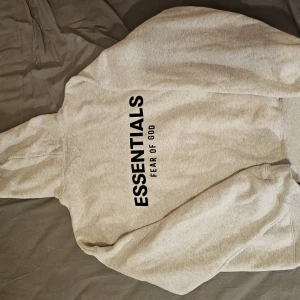 Grå Essentials Fear of God hoodie - Säljer en grå hoodie från Essentials Fear of God med svart tryck både fram och bak. Klassisk känguruficka, huva och lång ärm. Perfekt för dig som gillar streetwear och vill ha en clean look.