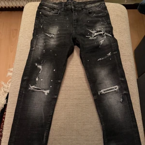 Svarta skinny jeans Supply & Demand - Svarta skinny jeans från Supply & Demand med slitningar och färgstänk för en edgy look. Jeansen har klassisk femficksdesign, dragkedja och knapp framtill. Snyggt tvättade detaljer och smal passform som sitter tajt längs benen.