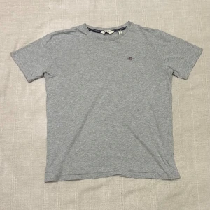 Grå t-shirt från GANT i bomull - Snygg grå t-shirt från GANT med klassisk passform och rund hals. T-shirten har ett litet broderat GANT-logo på bröstet och är tillverkad i mjuk bomull. Perfekt basic-plagg med stilren look.