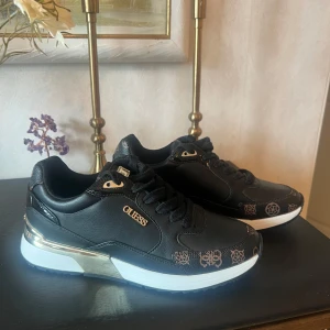 Svarta sneakers från Guess strl 39 - Snygga svarta sneakers från Guess med gulddetaljer och logomönster i brunt på tån och sidorna. Skorna har vit sula med metallic guldinslag och är tillverkade i syntetmaterial med snörning. Perfekta för dig som vill ha en trendig och sportig look. Nypris 1.500kr helt nya!