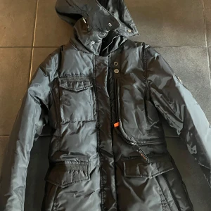 Svart dunjacka från Hollies, strl 36 - Svart lång dunjacka från Hollies med huva, flera fickor framtill och snygga detaljer som metallbadge på ärmen. Jackan har dragkedja och knappar framtill samt orange piping på insidan. Perfekt för kalla dagar när du vill hålla stilen. Visst, här är en annons för en Hollies-jacka:  perfekt skick! Den är värd runt 3800 kr, men jag säljer den för 1500 kr. Jackan är i absolut toppskick, inga skador eller hål, och har bara använts ett fåtal gånger. Den är supervarm, går att justera vid midjan 