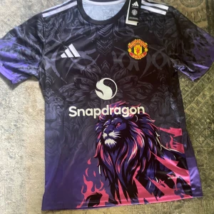Manchester United x Adidas lila fotbollströja - Unik Manchester United fotbollströja i lila och svart med stort lejonmotiv och detaljer i rosa och blått. Tröjan har korta ärmar, Adidas-logga, klubbmärke och Snapdragon som sponsor. Tillverkad i lätt och ventilerande material, perfekt för träning eller match.