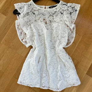 Vit spetstopp från Gina Tricot - Superfin vit topp från Gina Tricot med blommig spets och volangdetaljer vid axlarna. Den har rund hals och är kortärmad.