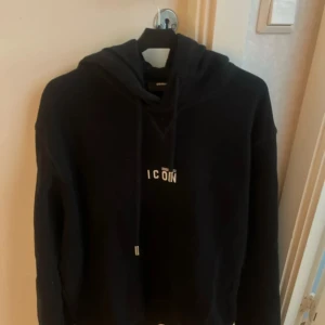 Svart hoodie från Dsquared2 ICON - Svart hoodie från Dsquared2 med vit 'ICON'-logga på bröstet. Klassisk modell med huva och snörning, tillverkad i mjuk bomull. Perfekt för dig som vill ha en stilren och trendig look med streetkänsla.