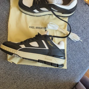 Axel Arigato svartvita sneakers - Snygga sneakers från Axel Arigato i svart och vitt skinn med beige detaljer. Klassisk siluett med perforerade partier, svart snörning och extra vita skosnören. Kommer med originalkartong och dustbag. Perfekta för dig som gillar stilrena och trendiga skor.