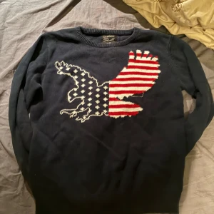 Mörkblå stickad tröja  - Mörkblå stickad tröja från Skill med en stor örn på bröstet i amerikanskt flaggmönster. Tröjan har rund halsringning och långa ärmar. Perfekt för dig som vill sticka ut med en cool och unik design. Perfekt för hösten! Storlek 146-152