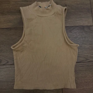 Beige ribbad polotopp ärmlös - Snygg beige polotopp i ribbad bomull med ärmlös design. Toppen har hög hals och croppad passform, perfekt att styla med jeans eller kjol för en trendig look.