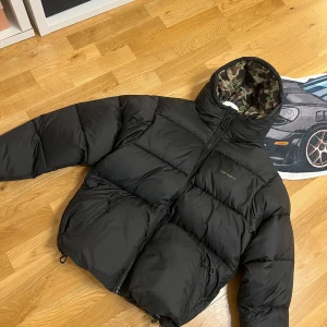 Svart pufferjacka Carhartt M - Säljer en svart pufferjacka från Carhartt i storlek M. Jackan har camouflagemönstrat foder i grönt och brunt, dragkedja framtill och två fickor med dragkedja. Perfekt för kalla dagar och riktigt snygg streetstil.
