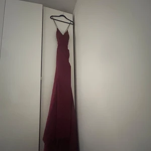 Vinröd långklänning med slits - Säljer en elegant vinröd långklänning med smala axelband och v-ringning. Klänningen har en figurnära passform och en snygg slits framtill. Perfekt för bal eller festliga tillfällen. Materialet är glansigt och faller snyggt längs kroppen.