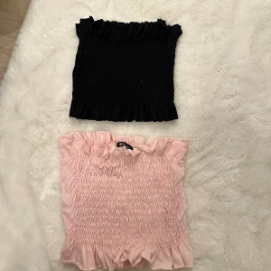 2 bandeau tubtoppar från Missguided - Två bandeau tubtoppar från Missguided i storlek 36. En är ljusrosa och en är svart, båda med smockad resår och volangkant upptill och nedtill. Perfekta att styla med jeans eller kjol för en trendig look.