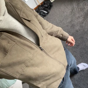 Grön mockajacka med dragkedja - Snygg grön mockajacka med dragkedja framtill och två bröstfickor med lock. Jackan har en klassisk krage och raka ärmar, perfekt för dig som gillar stilren och clean look. Passformen är normal och materialet känns mjukt och lyxigt. Nypris 1000kr