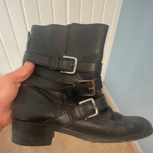 Svarta boots med spännen och klack - Snygga svarta boots i skinn med tre dekorativa spännen i silverfärg. Modellen har rund tå, låg klack och platt sula. Perfekta för dig som gillar en lite tuffare stil och vill ha något som funkar till många outfits.