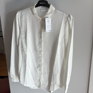 Vit klassisk blus från Vila - Snygg vit skjorta från Vila med klassisk krage och dolda knappar framtill. Skjortan har lång ärm och är tillverkad i ett mjukt, lätt material som ger en stilren look. Perfekt för dig som gillar en clean och enkel stil. Helt ny med lapp kvar ord pris 399 