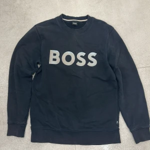 Svart sweatshirt från BOSS - Svart sweatshirt från BOSS med stor silverfärgad logga på bröstet. Klassisk rund halsringning och långa ärmar. Tillverkad i mjuk bomull för en bekväm känsla. Perfekt för en stilren och avslappnad look.