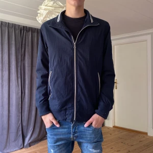 Marinblå vindjacka från Bison - Snygg marinblå vindjacka från Bison med vita detaljer på dragkedjor och krage. Jackan har två fickor med dragkedja, ribbade muddar och sportig känsla. Perfekt för vår och höst när du vill ha en stilren look. Ny pris 1800kr men det låga priset eftersom jag måste rensa min garderob