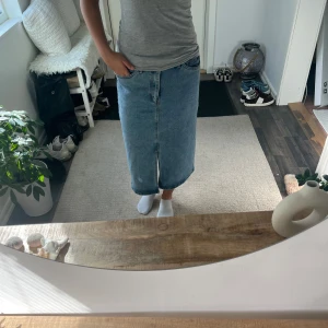 Blå jeanskjol med slits - Säljer en klassisk blå lång jeanskjol med rak passform och slits framtill. Kjolen har fem fickor, knapp och dragkedja i midjan samt bälteshällor. Perfekt för dig som gillar enkel och tidlös denimstil.