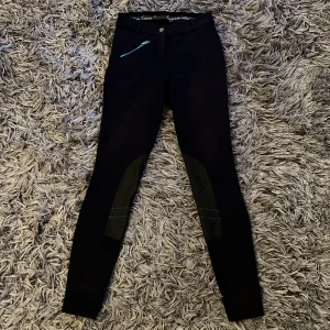 Svarta ridbyxor från Fouganza - Svarta ridbyxor från Fouganza med snygga detaljer och en turkos dragkedja vid fickan. På insidan av benen finns extra grepp och komfort vid ridning. Tight passform och midja med knapp och dragkedja.  Storlek 8-11 åringar 🐴