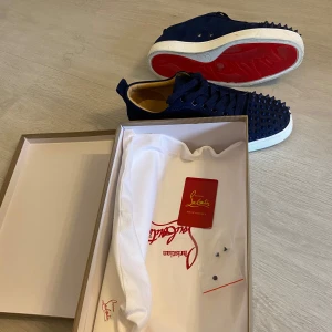 Blå mocka sneakers från Louboutin - Säljer ett par mörkblå sneakers från Christian Louboutin i mocka med ikoniska röda sulor. Skorna har snörning och coola nitar över tån för en edgy look. Insidan är beige och sulan är vit. Kommer med originalkartong och dustbag.