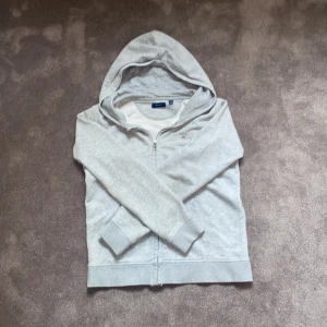 Grå zip hoodie från GANT - Säljer en stilren grå hoodie från GANT med dragkedja framtill och broderad logga på bröstet. Tröjan har huva, känguruficka och är tillverkad i mjuk bomullsmix. Perfekt för chill dagar eller när du vill ha en enkel och snygg look. Hör av dig vid minsta fundering! Priset är prutbart