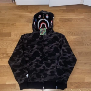 BAPE Shark Camo Hoodie svart/grå - Säljer en ikonisk BAPE Shark Hoodie i svart och grå camo med dragkedja hela vägen och huva med hajansikte och broderade detaljer. Klassisk BAPE-logga på ärmslutet och färgglada bokstäver på huvan. Perfekt statement-plagg för streetwear.