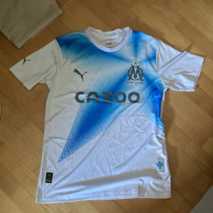 Marseille fotbollströja Puma vit/blå - Olympique de Marseille matchtröja från Puma i vitt med blått grafiskt mönster över bröstet. Tryckt klubbmärke, sponsorlogga och Puma-loggor på ärmarna. Tillverkad i lätt och ventilerande material, perfekt för fotboll eller supporterliv.