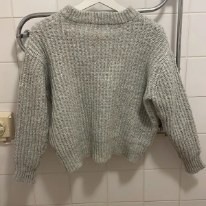 Grå stickad tröja med grov struktur - Säljer en grå stickad tröja med grov struktur och ribbade muddar. Tröjan har en avslappnad passform och hög hals, perfekt för kyliga dagar. Enkel att matcha med jeans eller kjol 