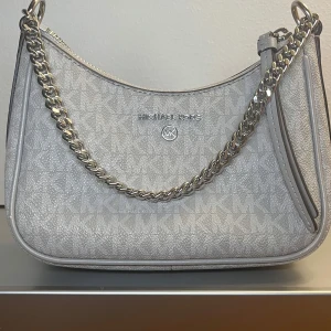 Grå axelväska Michael Kors med kedja - Snygg axelväska från Michael Kors i ljusgrått med MK-logga över hela väskan. Väskan har en silvrig kedja som axelrem och dragkedja upptill. Tillverkad i syntetmaterial med detaljer i skinn. Perfekt storlek för det viktigaste och ger en clean look.