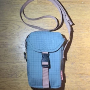 Grå axelväska från Comme des Garçons - Snygg liten axelväska i grått med rutigt mönster och beige justerbar rem. Väskan har ett spänne framtill och en praktisk ficka på baksidan. Perfekt för mobilen och småprylar. Märkt med Comme des Garçons på sidan.