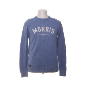 Blå stickad tröja från Morris Gents - Snygg blå stickad tröja från Morris Gents med vit text 'Morris Company' på bröstet. Tröjan har rund halsringning, ribbade muddar och är långärmad. Perfekt för dig som gillar klassisk och stilren look.