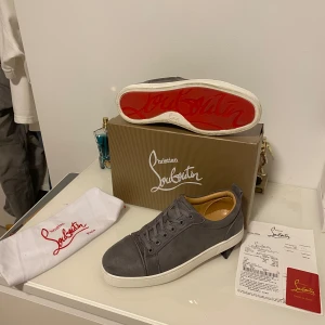 Grå mocka sneakers från Christian Louboutin - Säljer ett par gråa sneakers från Christian Louboutin i lyxig mocka med klassisk röd sula. Skorna har rund tå, snörning och vit platt sula. Insidan är i beige skinn och de levereras med originalkartong, dustbag och äkthetskort.