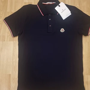 Marinblå Moncler pikétröja med logga - Snygg marinblå pikétröja från Moncler med klassisk krage och knäppning framtill. Tröjan har Moncler-logga på bröstet och röda/vita detaljer på krage och ärmslut. Tillverkad i bomull för en skön känsla och stilren look.