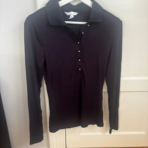 Svart långärmad topp med knappar - Stilren svart långärmad polotopp med klassisk krage och guldiga knappar framtill. Toppen är ribbad och har en slim passform som sitter snyggt på kroppen. Perfekt för dig som gillar en enkel men ändå elegant look. Helt ny, aldrig använd. Säljer då jag råkade riva av prislappen. 