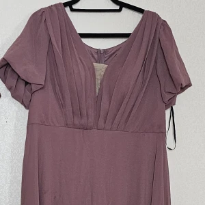 Ljuslila långklänning med v-ringning - Elegant ljuslila långklänning med djup v-ringning och veckade detaljer upptill. Klänningen har korta, lösa ärmar och dragkedja bak. Perfekt för dig som vill ha en flowy och stilren look.