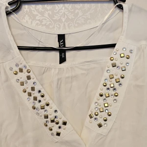 Vit blus med pärlor och nitar från VIX - Vit blus från VIX med v-ringning dekorerad med pärlor, guldiga nitar och glittriga stenar. Långärmad modell med knappar framtill och lätt rynkade ärmslut. Tunt, mjukt tyg som ger en luftig känsla. Perfekt för dig som gillar detaljer och en chic look.