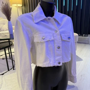 Vit croppad jeansjacka med knappar - Snygg vit croppad jeansjacka med rå kant och silverfärgade knappar. Jackan har två bröstfickor med lock och klassisk krage. Perfekt för dig som vill ha en trendig och avslappnad look. Passar perfekt i S/M storlek..
