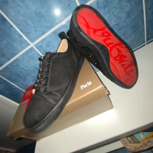Svarta sneakers från Christian Louboutin - Säljer ett par svarta sneakers från Christian Louboutin med klassisk röd sula och diskret logga. Skorna har snörning, ovandel i mocka och insida i ljust skinn. Perfekta för dig som vill ha en lyxig och stilren look.
