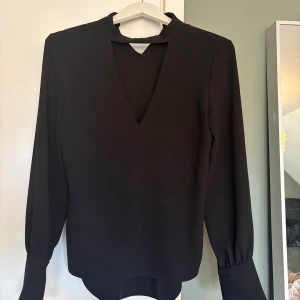 Svart blus med v-ringning - Stilren svart blus med lång ärm och markerad v-ringning framtill. Blusen har en hög krage med öppning och lätt puffade ärmslut. Perfekt för dig som gillar en enkel men trendig look.