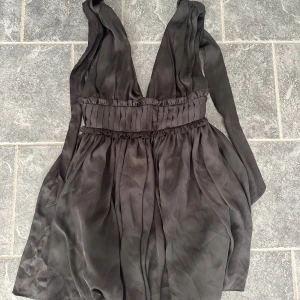 Svart omlottklänning med knyt - Supersnygg svart omlottklänning med djup v-ringning och bred midjedetalj. Klänningen har knytband vid axlarna och är tillverkad i ett glansigt, mjukt material som ger en lyxig känsla. Perfekt för dig som vill sticka ut med en elegant och trendig look.