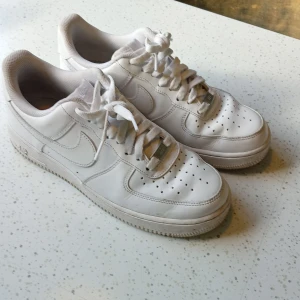 Nike Air Force 1 vita sneakers - Klassiska Nike Air Force 1 sneakers i helvitt skinn med perforerad tå och rundad sula. Lågt skaft, snörning och ikonisk Swoosh-logga på sidan. Perfekt för dig som gillar clean och tidlös streetstil.